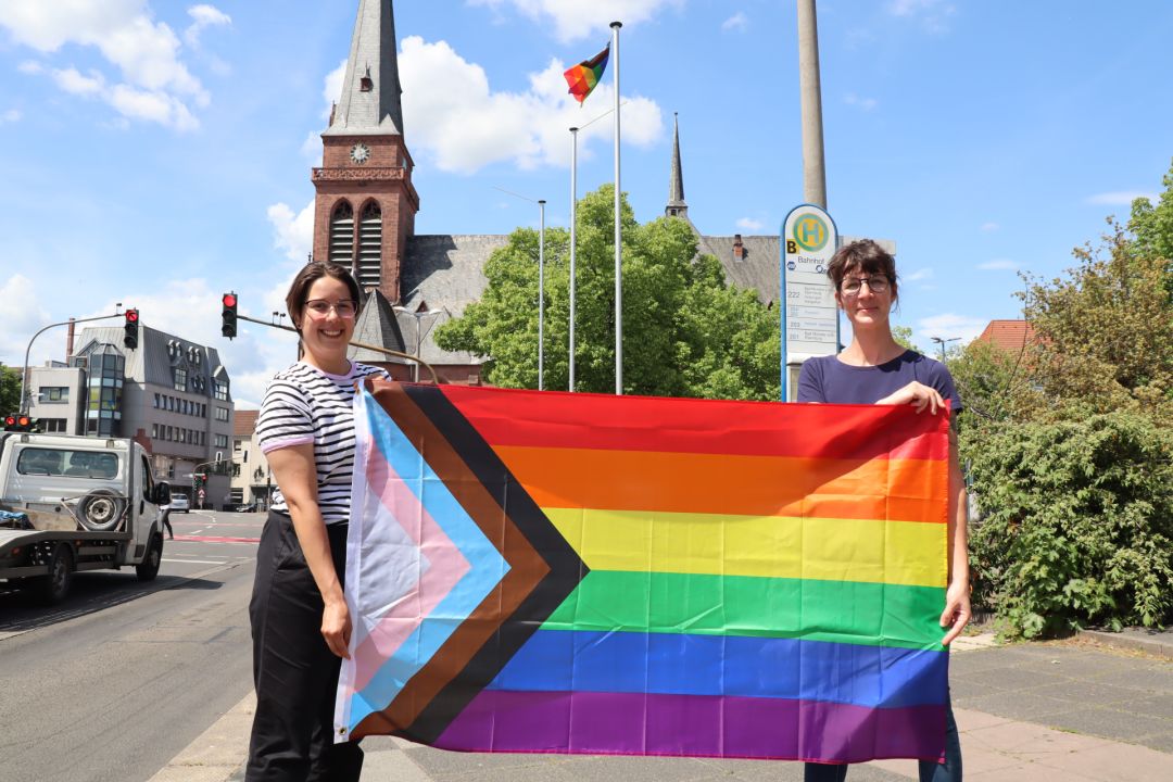 Laura Schäfer und Danielle Schulz halten gemeinsam die Pride Flag am Europaplatz, im Hintergrund die gehisste Regenbogenfahne. Und die Kirche Heilig Kreuz. :)