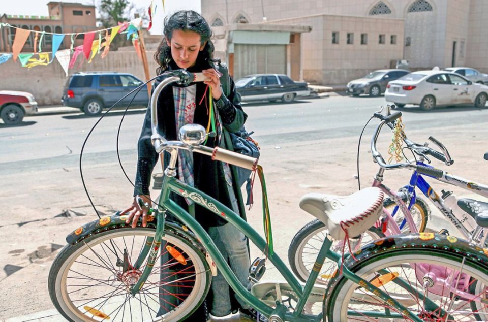 Szene aus dem Film: Eine Frau steht hinter einem Fahrrad und betrachtet es prüfend. Im Hintergrund eine befahrene Straße mit Autos.
